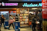 A busy newsstand with various magazines and newspapers on display. The scene includes people walking and browsing, with a Dunkin' Donuts and a Coca-Cola vending machine visible. Bright signage and numerous publications create a bustling and vibrant environment.