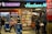 A busy newsstand with various magazines and newspapers on display. The scene includes people walking and browsing, with a Dunkin' Donuts and a Coca-Cola vending machine visible. Bright signage and numerous publications create a bustling and vibrant environment.