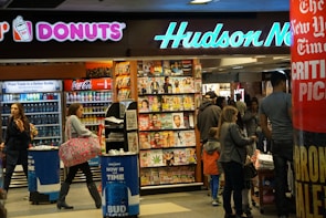 A busy newsstand with various magazines and newspapers on display. The scene includes people walking and browsing, with a Dunkin' Donuts and a Coca-Cola vending machine visible. Bright signage and numerous publications create a bustling and vibrant environment.