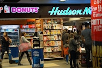 A busy newsstand with various magazines and newspapers on display. The scene includes people walking and browsing, with a Dunkin' Donuts and a Coca-Cola vending machine visible. Bright signage and numerous publications create a bustling and vibrant environment.