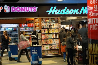 A busy newsstand with various magazines and newspapers on display. The scene includes people walking and browsing, with a Dunkin' Donuts and a Coca-Cola vending machine visible. Bright signage and numerous publications create a bustling and vibrant environment.