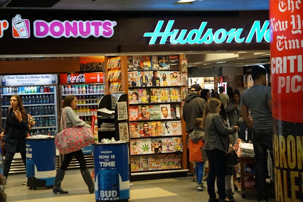 A busy newsstand with various magazines and newspapers on display. The scene includes people walking and browsing, with a Dunkin' Donuts and a Coca-Cola vending machine visible. Bright signage and numerous publications create a bustling and vibrant environment.
