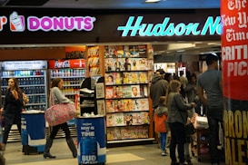 A busy newsstand with various magazines and newspapers on display. The scene includes people walking and browsing, with a Dunkin' Donuts and a Coca-Cola vending machine visible. Bright signage and numerous publications create a bustling and vibrant environment.