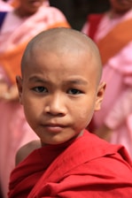A young person wearing a red robe stands in focus, with a serious expression and shaved head. Others in similar colorful attire are blurred in the background, suggesting a group or community setting.