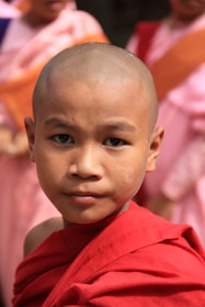 A young person wearing a red robe stands in focus, with a serious expression and shaved head. Others in similar colorful attire are blurred in the background, suggesting a group or community setting.