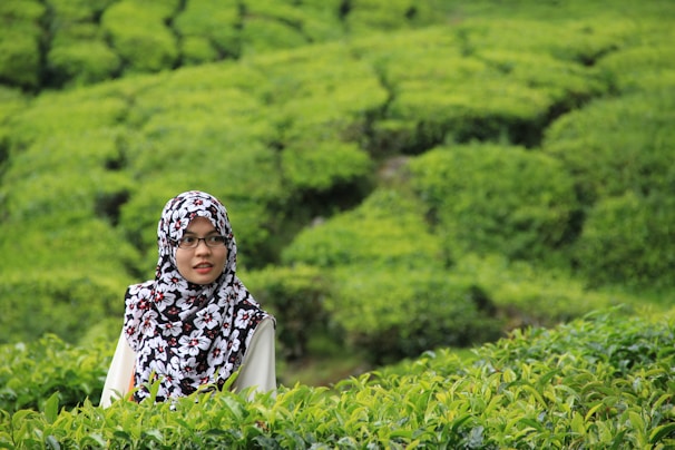 A woman in a floral patterned hijab stands amidst lush green tea plantations. The landscape features well-maintained rows of tea plants that create a textured, verdant backdrop.