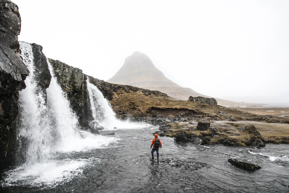 man with backpack standing on river beside waterfalls