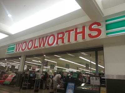 A storefront with a large red and white sign that reads 'Woolworths'. Below the sign is a glass entrance to a supermarket where several people are shopping. Various checkout counters are visible, along with aisles stocked with products.