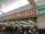 A storefront with a large red and white sign that reads 'Woolworths'. Below the sign is a glass entrance to a supermarket where several people are shopping. Various checkout counters are visible, along with aisles stocked with products.