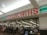 A storefront with a large red and white sign that reads 'Woolworths'. Below the sign is a glass entrance to a supermarket where several people are shopping. Various checkout counters are visible, along with aisles stocked with products.