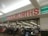 A storefront with a large red and white sign that reads 'Woolworths'. Below the sign is a glass entrance to a supermarket where several people are shopping. Various checkout counters are visible, along with aisles stocked with products.