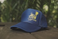 A navy blue baseball cap with a yellow graphic design featuring a house and palm tree. The cap is placed on a wooden surface with a blurred natural green background.
