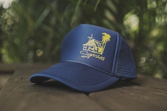 A navy blue baseball cap with a yellow graphic design featuring a house and palm tree. The cap is placed on a wooden surface with a blurred natural green background.