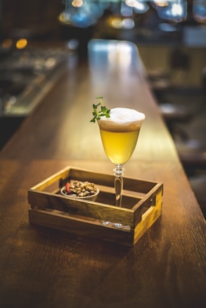 A cocktail glass containing a yellow drink with frothy cream on top is elegantly placed on a wooden table. A small garnish adds a touch of green to the visually appealing drink. Next to it, in a wooden tray, sits a small bowl filled with nuts and a red chili pepper. The background of the scene is a softly blurred bar setting, creating a warm and inviting atmosphere.