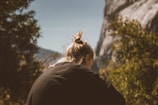A person with a blonde hair bun, wearing a black shirt, appears to be enjoying a natural outdoor setting. The background features a rocky mountain and lush greenery on a sunny day, creating a serene and peaceful atmosphere.