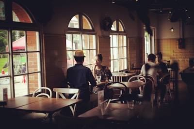 A cozy cafe environment with people sitting at tables, engaged in conversation. Large windows allow natural light to filter in, creating a warm and inviting atmosphere. The interior features wooden tables and a mix of white and dark chairs, with subtle decor enhancing the relaxed vibe.