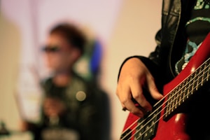 A close-up view of a person's hand playing a red electric bass guitar, with blurred background elements including a person wearing sunglasses and a leather jacket.