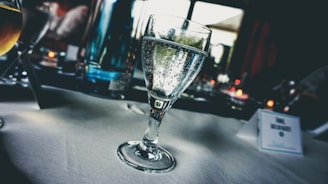 A glass of sparkling water sits on a white tablecloth with bubbles visible inside the glass. The background has a blurred view of a table setting, including other glassware and a softly focused outside view through a window, suggesting a relaxed dining environment.