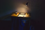 A warm wooden desk with a vintage lamp, laptop, and scattered planner pages.