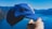 A hand is holding a blue baseball cap with a white logo in front of a scenic background of a lake and mountains under a clear blue sky.