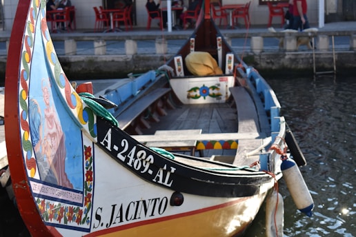 A traditional wooden boat, decorated with colorful paintings, is docked along a canal. The front panel of the boat features a vibrant illustration surrounded by a floral pattern. The name 'S. Jacinto' and a registration number are displayed along the side. In the background, there are red tables and chairs outside a building, suggesting a café or dining area by the water.