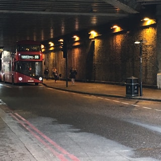 A red double-decker bus is traveling through a dimly lit tunnel with yellow-orange lights lining the walls. There are several pedestrians walking along the sidewalk beside the bus.