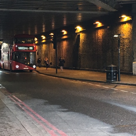 A red double-decker bus is traveling through a dimly lit tunnel with yellow-orange lights lining the walls. There are several pedestrians walking along the sidewalk beside the bus.