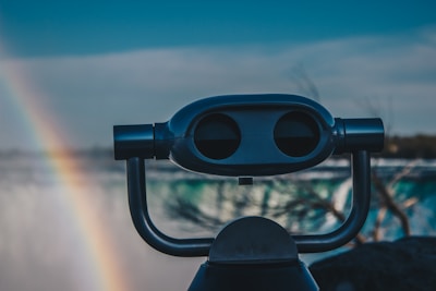 A pair of stationary binoculars overlooks a scenic view, with a rainbow visible in the background against a blurred natural landscape.