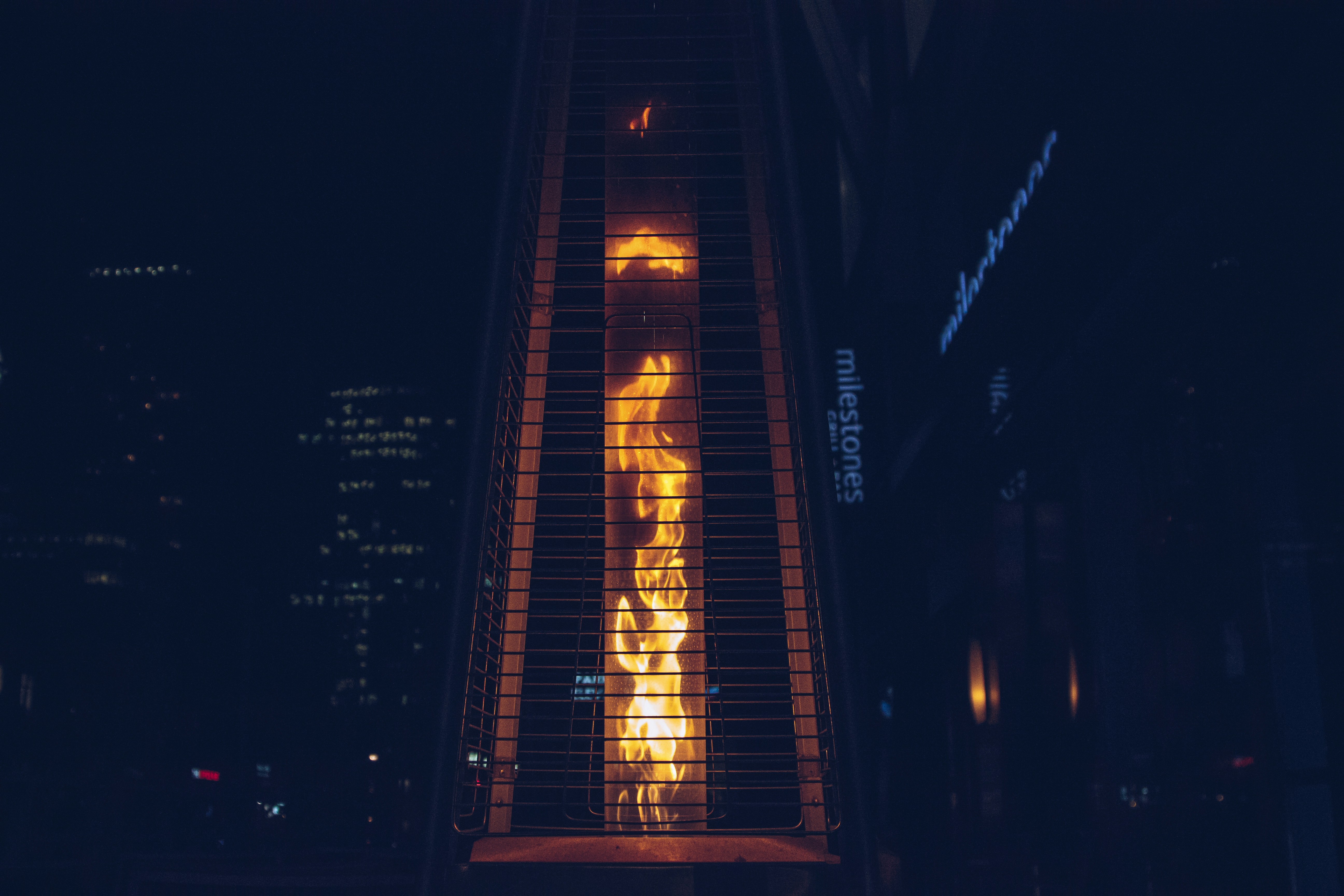 A tall, outdoor gas heater with visible flames is enclosed within a protective metal casing. The background is a dark urban setting with dimly lit buildings and a sign partially visible.