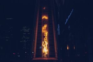 A tall, outdoor gas heater with visible flames is enclosed within a protective metal casing. The background is a dark urban setting with dimly lit buildings and a sign partially visible.