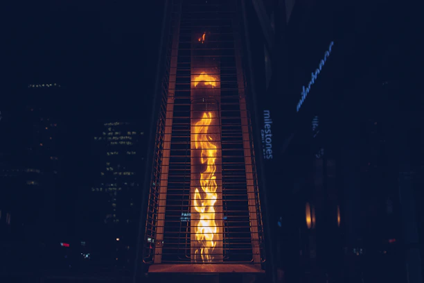 A tall, outdoor gas heater with visible flames is enclosed within a protective metal casing. The background is a dark urban setting with dimly lit buildings and a sign partially visible.