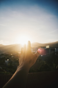 A hand reaching towards the bright sun, with rays of sunlight illuminating the scene. The background contains soft-focus elements of a landscape with hills and possibly some buildings. The colors create a warm and hopeful ambiance.