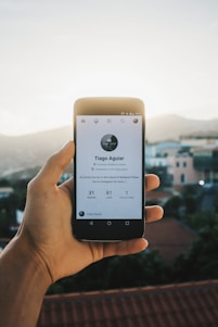 A hand is holding a smartphone displaying a social media profile. The profile features a minimalistic design with a profile picture, name, and some statistics below it, set against a blurred outdoor background with houses and a clear sky.