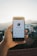 A hand is holding a smartphone displaying a social media profile. The profile features a minimalistic design with a profile picture, name, and some statistics below it, set against a blurred outdoor background with houses and a clear sky.