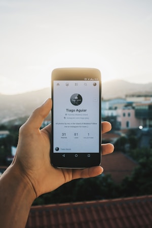 A hand is holding a smartphone displaying a social media profile. The profile features a minimalistic design with a profile picture, name, and some statistics below it, set against a blurred outdoor background with houses and a clear sky.
