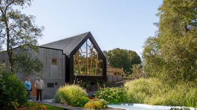 A modern house with a sharp, angular roof and large glass windows is surrounded by lush greenery, including trees and a well-maintained garden. Two people stand near the entrance, enjoying the serene setting.
