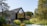 A modern house with a sharp, angular roof and large glass windows is surrounded by lush greenery, including trees and a well-maintained garden. Two people stand near the entrance, enjoying the serene setting.