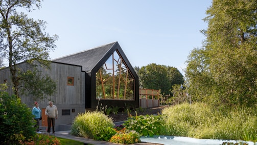 A modern house with a sharp, angular roof and large glass windows is surrounded by lush greenery, including trees and a well-maintained garden. Two people stand near the entrance, enjoying the serene setting.