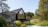 A modern house with a sharp, angular roof and large glass windows is surrounded by lush greenery, including trees and a well-maintained garden. Two people stand near the entrance, enjoying the serene setting.