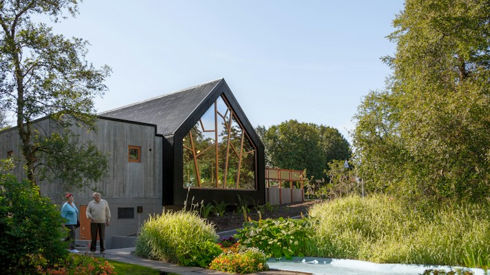A modern house with a sharp, angular roof and large glass windows is surrounded by lush greenery, including trees and a well-maintained garden. Two people stand near the entrance, enjoying the serene setting.