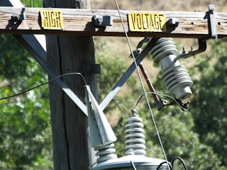 A wooden utility pole featuring transformers and insulators with a 'High Voltage' warning sign. The background is blurred with greenery, indicating a natural outdoor setting.
