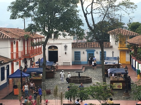 A small, quaint plaza with traditional colonial-style architecture, featuring white buildings with colorful accents. There's a central fountain surrounded by cobblestone pavement. Several stalls with blue canopies are set up, likely selling local goods or crafts. People are walking and shopping in the area, some dressed casually while a few are in white attire.