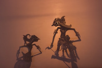 Two intricate shadow puppets are cast on a warm, softly lit background. The figures are detailed with elaborate headdresses and ornate costumes, conveying a traditional or cultural aesthetic.