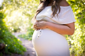A pregnant woman in a white dress stands in a sunlit outdoor environment surrounded by lush greenery. Her hands rest gently on her belly, and she wears a ring on her left hand.