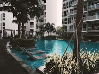 A serene outdoor pool area surrounded by modern residential buildings and lush greenery. The scene includes several lounge chairs partially submerged in the water, palm trees, and is bathed in natural light.