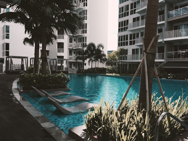 A serene outdoor pool area surrounded by modern residential buildings and lush greenery. The scene includes several lounge chairs partially submerged in the water, palm trees, and is bathed in natural light.