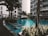 A serene outdoor pool area surrounded by modern residential buildings and lush greenery. The scene includes several lounge chairs partially submerged in the water, palm trees, and is bathed in natural light.