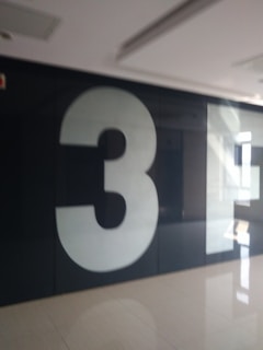 A large, white number three is prominently displayed on a dark wall in a hallway with a glossy floor reflecting light from windows.