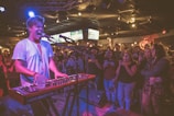 A musician passionately performs on stage, playing a keyboard and singing into a microphone. The performer is wearing a white shirt and a necklace, with vibrant stage lights casting a mix of colors. An enthusiastic audience in casual attire surrounds the stage, some taking photos and videos with their phones. The venue has a lively atmosphere, with screens visible in the background displaying sports.