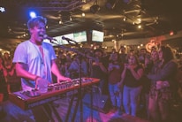 A musician passionately performs on stage, playing a keyboard and singing into a microphone. The performer is wearing a white shirt and a necklace, with vibrant stage lights casting a mix of colors. An enthusiastic audience in casual attire surrounds the stage, some taking photos and videos with their phones. The venue has a lively atmosphere, with screens visible in the background displaying sports.